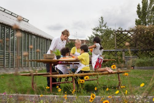 Eine Köchin schneidet zusammen mit 4 Kindern draußen auf einer Garten-Bank Gemüse