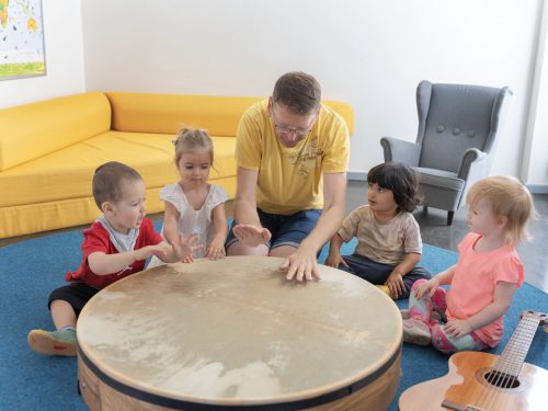 Ein Erzieher trommelt zusammen mit Kita-Kindern auf einer großen flachen Trommel, daneben liegt eine Gitarre